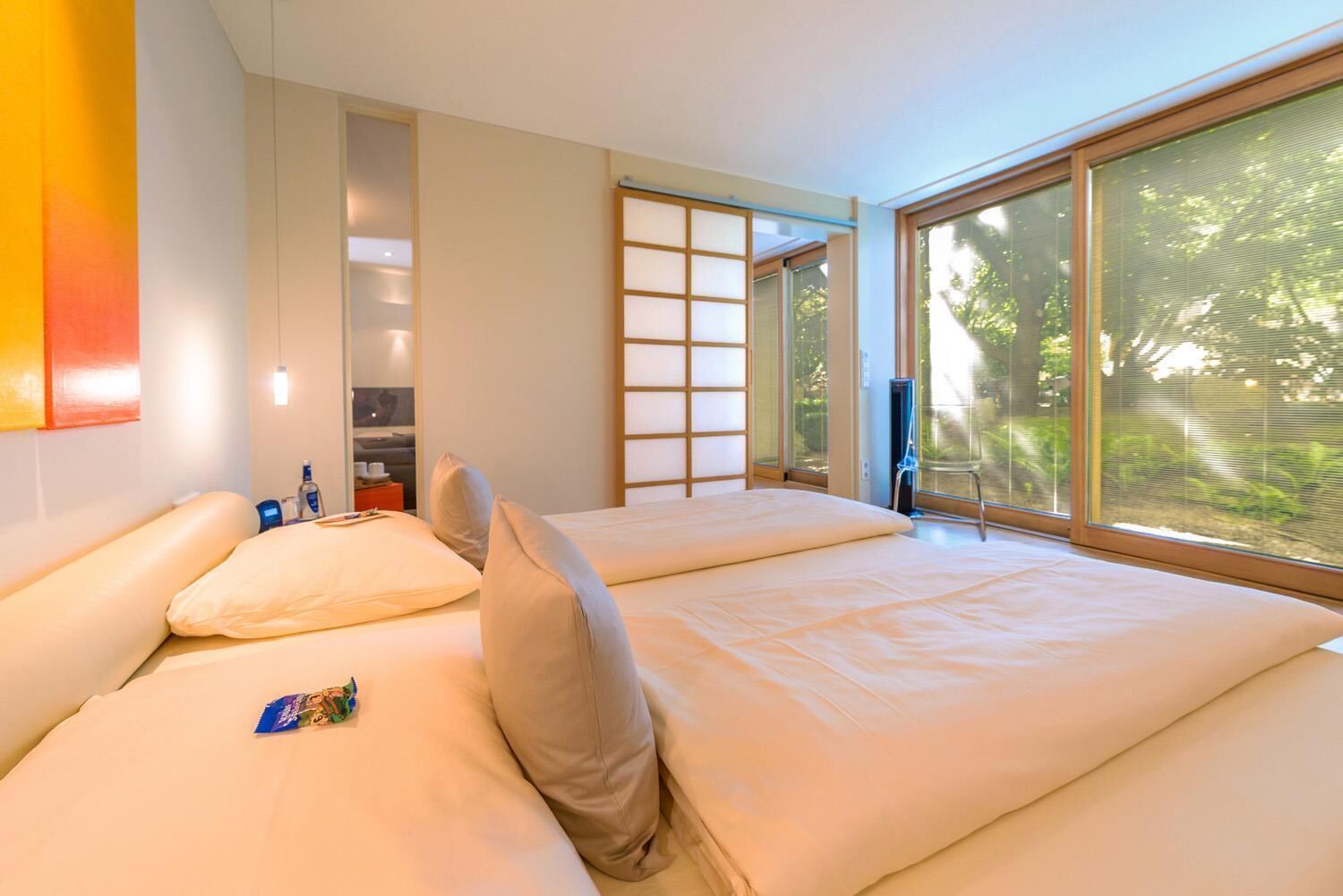 A modern hotel room with a large bed, a small table with a bottle and glasses, and a view of greenery through large windows.