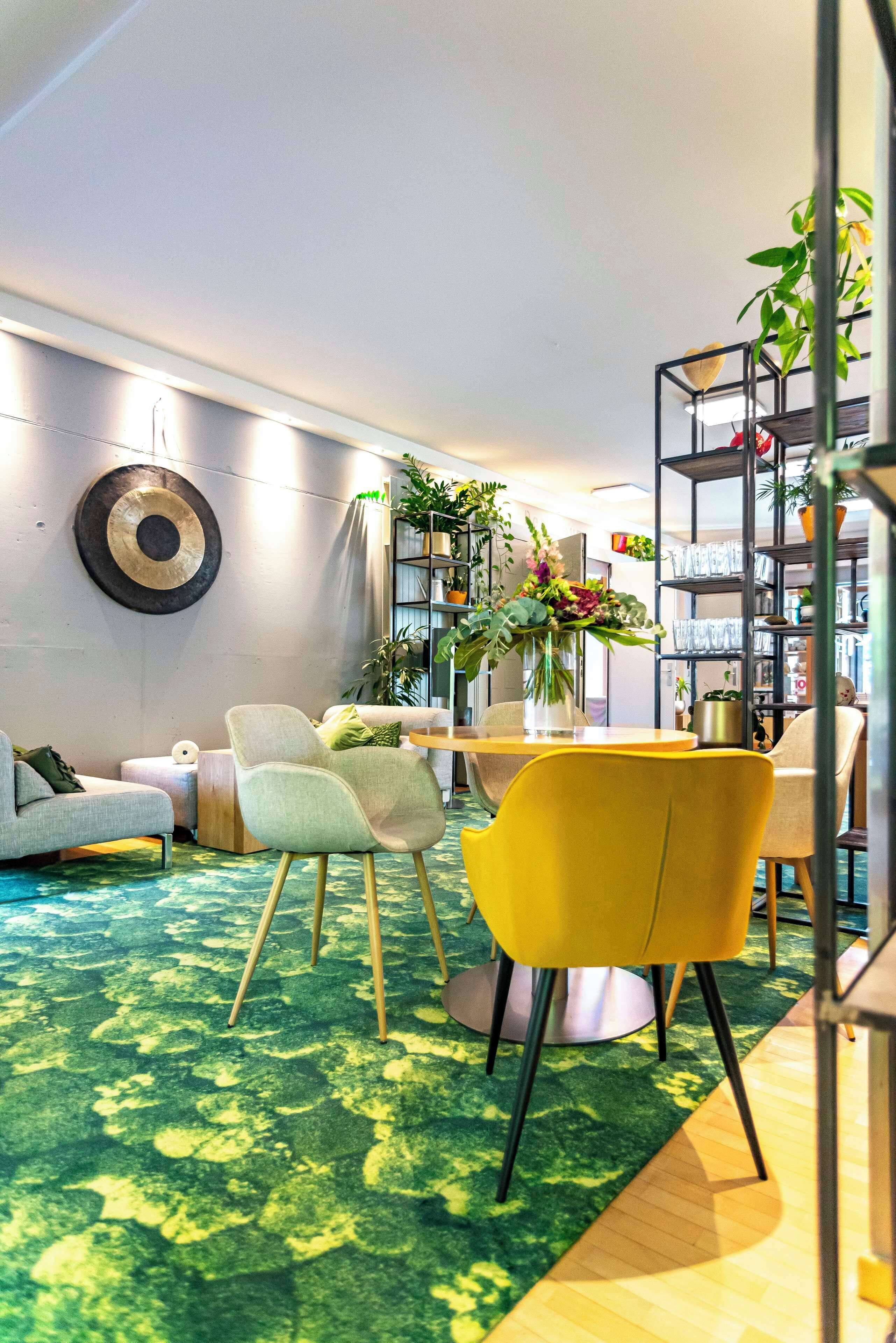A modern living room with a green carpet, a yellow chair, a grey sofa, and a bookshelf filled with plants and books.