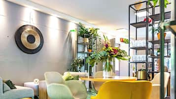 A modern living room with a green carpet, a yellow chair, a grey sofa, and a bookshelf filled with plants and books.