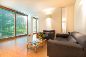 A modern living room with a sofa, armchair, and coffee table. There is a large window with blinds, a wall-mounted light, and a view of greenery outside.