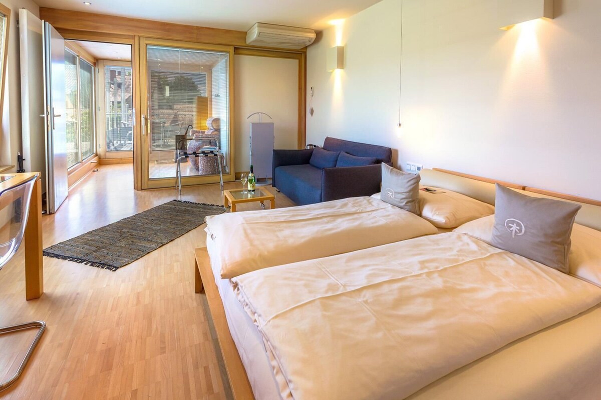 Una habitación de hotel moderna con una cama grande, un sofá, una mesita y una puerta de cristal que da a un balcón.