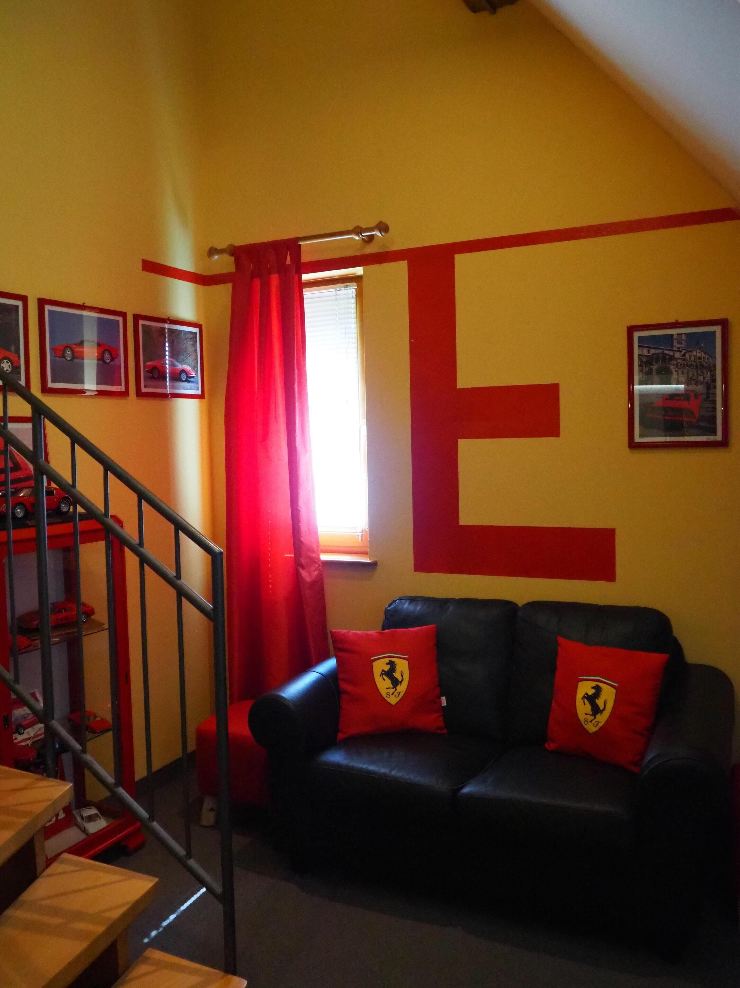 Um quarto com uma grande letra 'E' na parede, um sofá de couro preto com almofadas vermelhas, uma escada e uma janela com cortinas vermelhas.