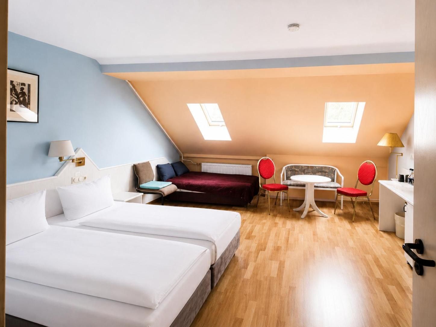 Ein Hotelzimmer mit einem großen Bett, einem Schreibtisch, einem Sessel und einem kleinen Tisch. Zwei Fenster sorgen für natürliches Licht.