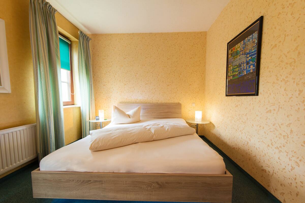 A hotel room with a wooden bed, two bedside lamps, a window with curtains, a radiator, and a framed picture on the wall.