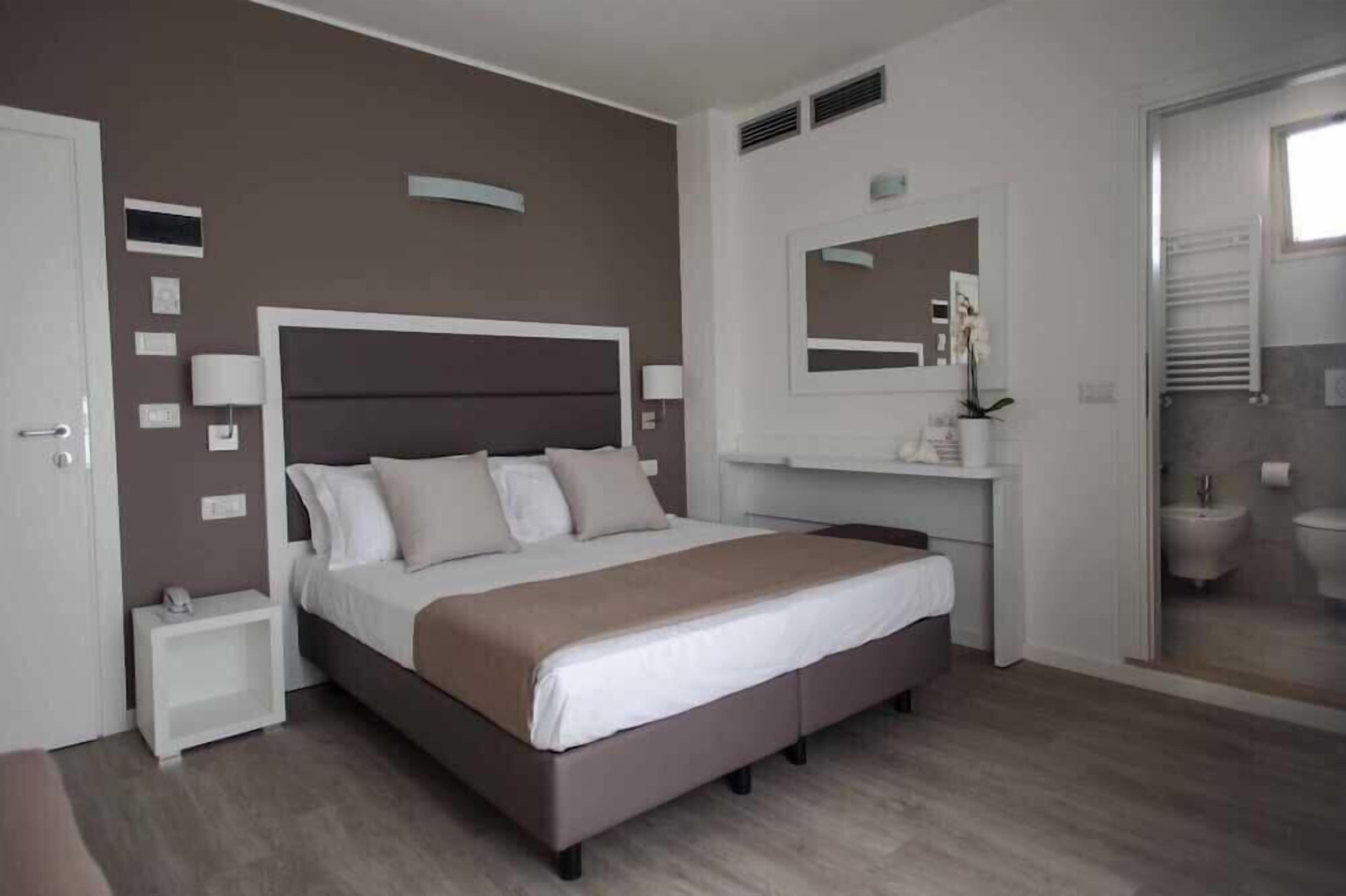 A modern bedroom with a large bed, bedside tables, a mirror, and a bathroom visible through a glass door.