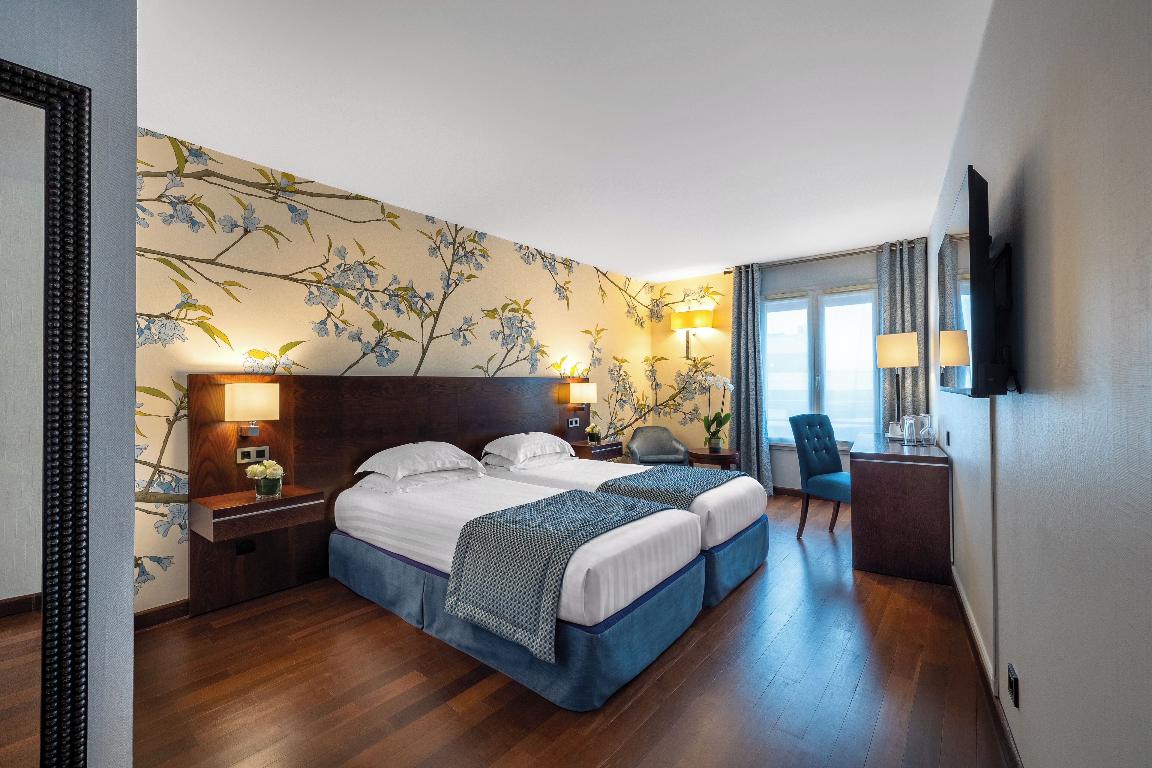 A hotel room with two beds, a desk, a chair, a TV, and a window with curtains.