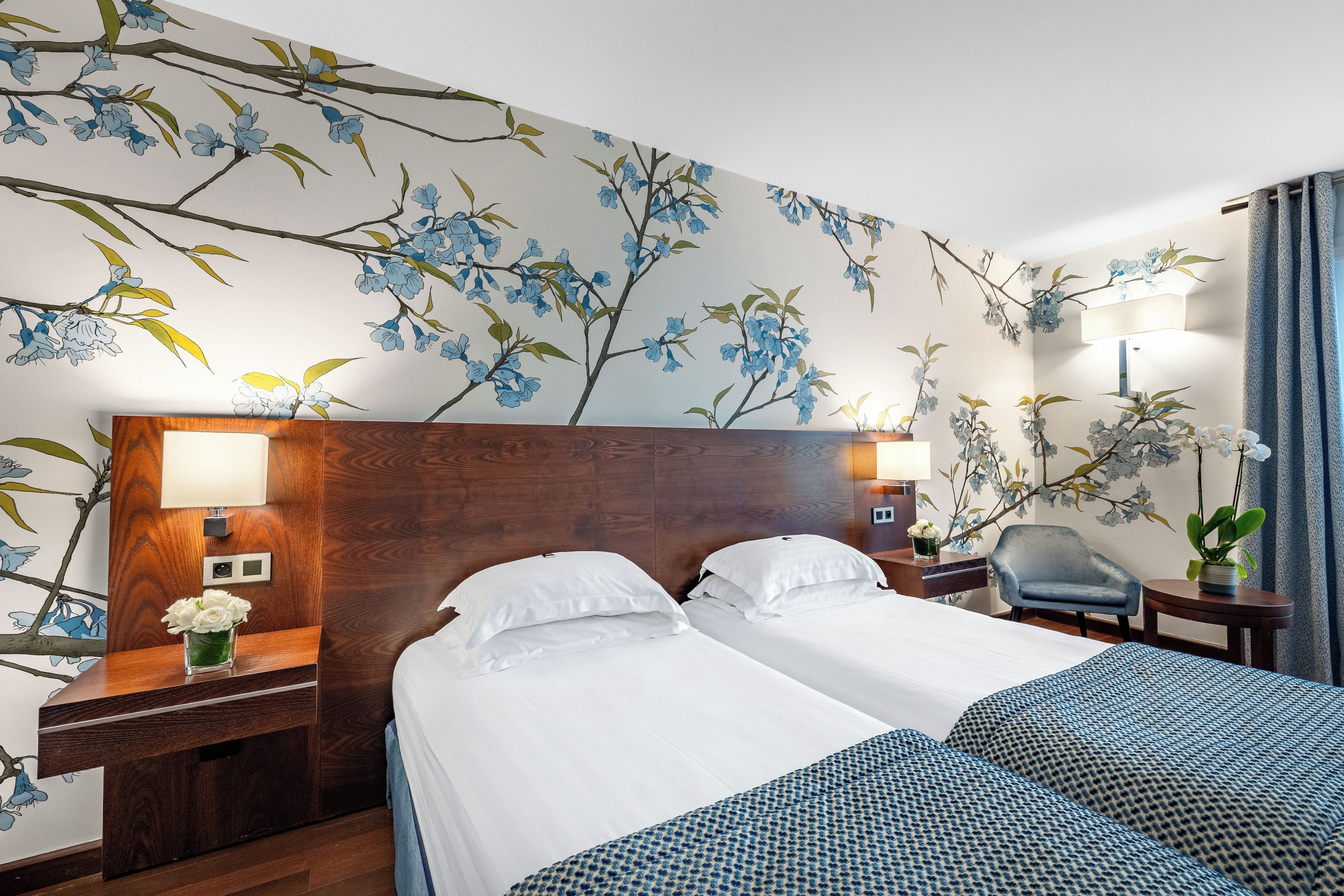 A hotel room with a large bed, a nightstand, a lamp, a chair, and a wall with a floral pattern.
