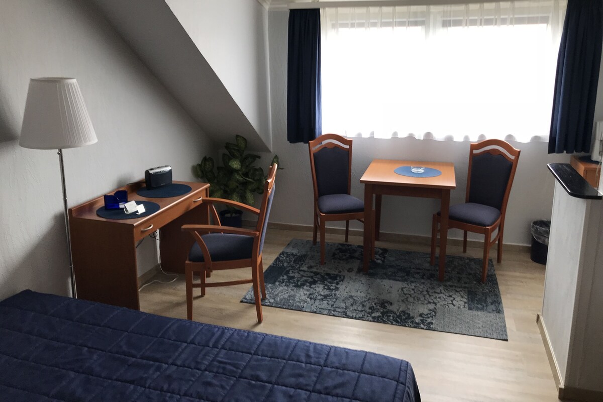 A hotel room with a bed, a desk with a lamp, two chairs, a small table, and a window with curtains.