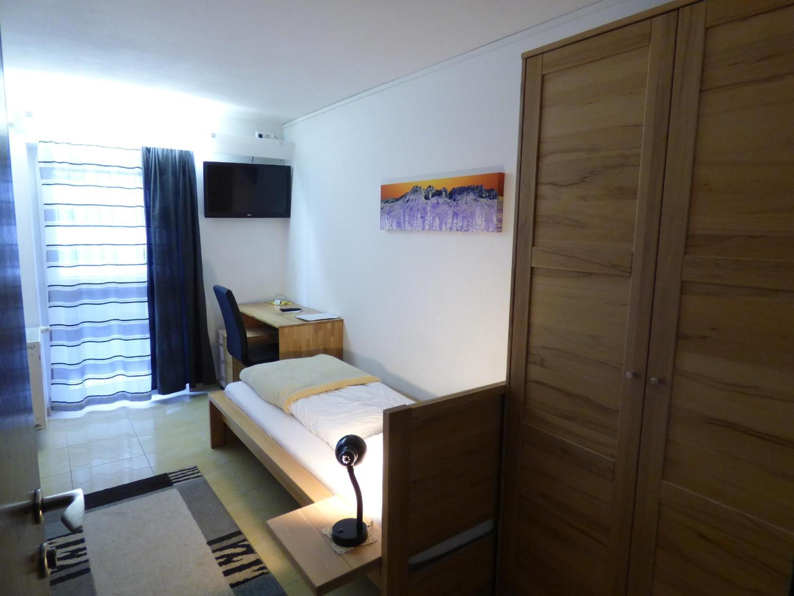 A small, well-lit room with a single bed, a desk, and a chair. A television is mounted on the wall, and a painting hangs above the desk.