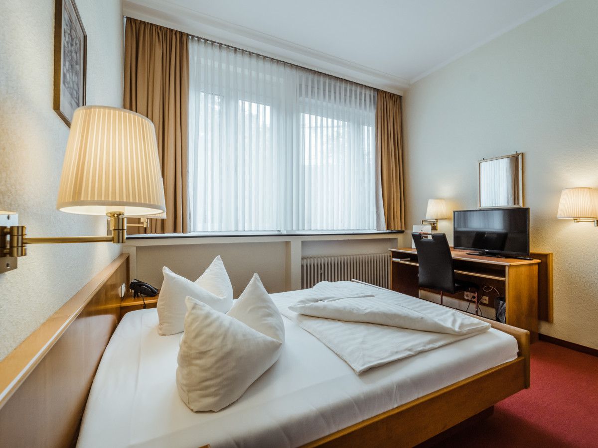A hotel room with a bed, a desk with a television, a chair, a lamp, and a window with curtains.