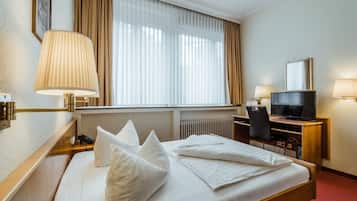 A hotel room with a bed, a desk with a television, a chair, a lamp, and a window with curtains.