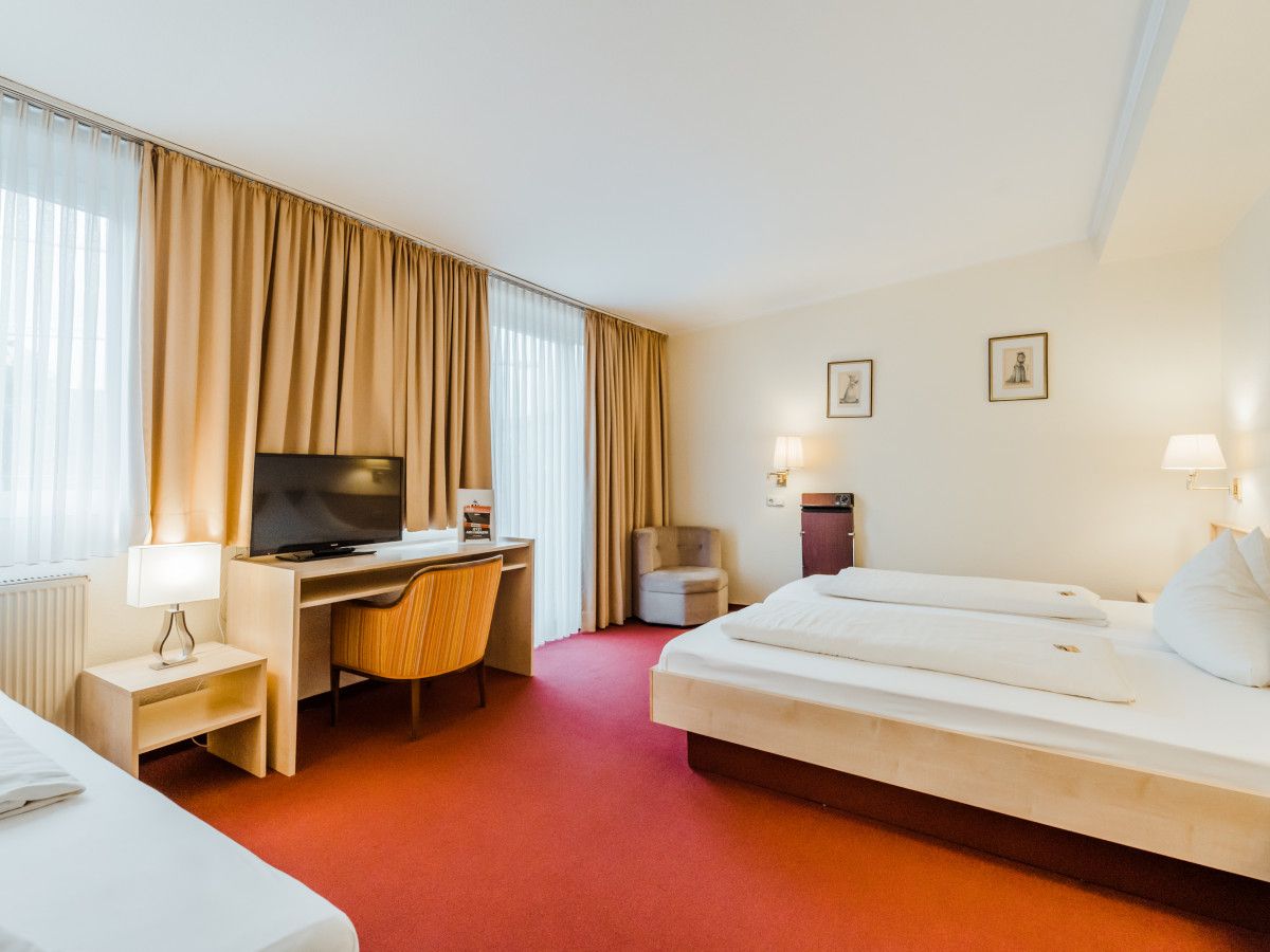 Habitación de hotel con cama, escritorio con televisor, una silla, una mesita con lámpara y ventana con cortinas.
