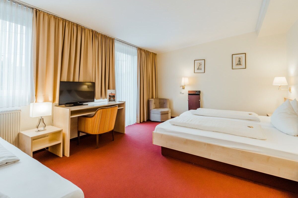 A hotel room with a bed, a desk with a television, a chair, a small table with a lamp, and a window with curtains.
