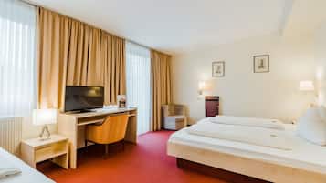 Habitación de hotel con cama, escritorio con televisor, una silla, una mesita con lámpara y ventana con cortinas.
