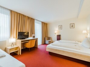 A hotel room with a bed, a desk with a television, a chair, a small table with a lamp, and a window with curtains. - Hotel Am Marschiertor (Aachen)