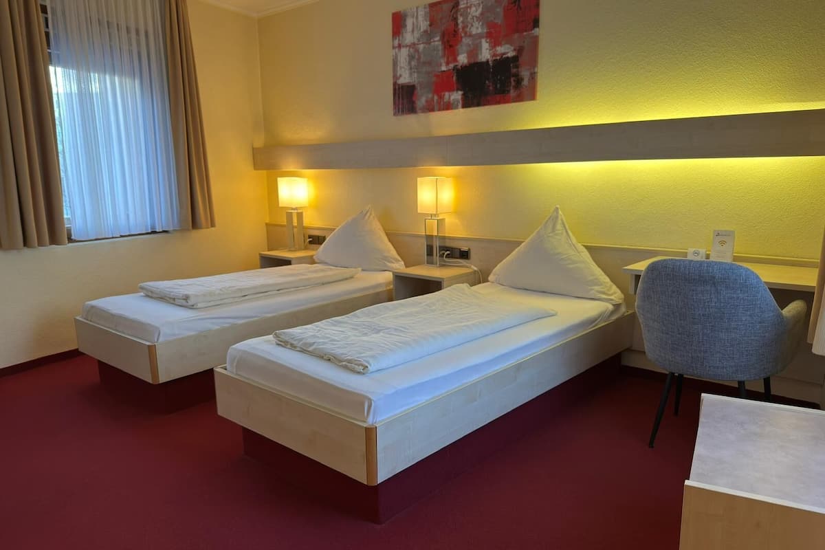 A hotel room with two single beds, a desk, a chair, and a window with curtains.