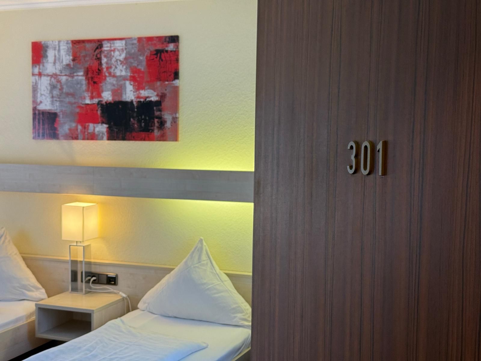 A hotel room with two beds, a bedside table with a lamp, a wall-mounted artwork, and a door with the number 301.