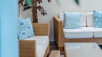 A wicker sofa with white cushions and patterned pillows, a potted plant, and a textured rug.