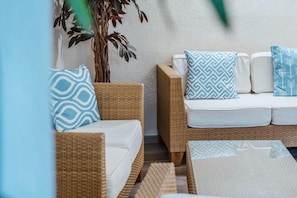 A wicker sofa with white cushions and patterned pillows, a potted plant, and a textured rug. - Hotel Am Marschiertor (Aachen)