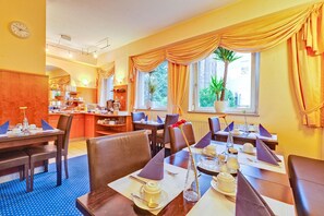 Un restaurante con mesas dispuestas para comer, una zona de cocina y grandes ventanales con cortinas.
