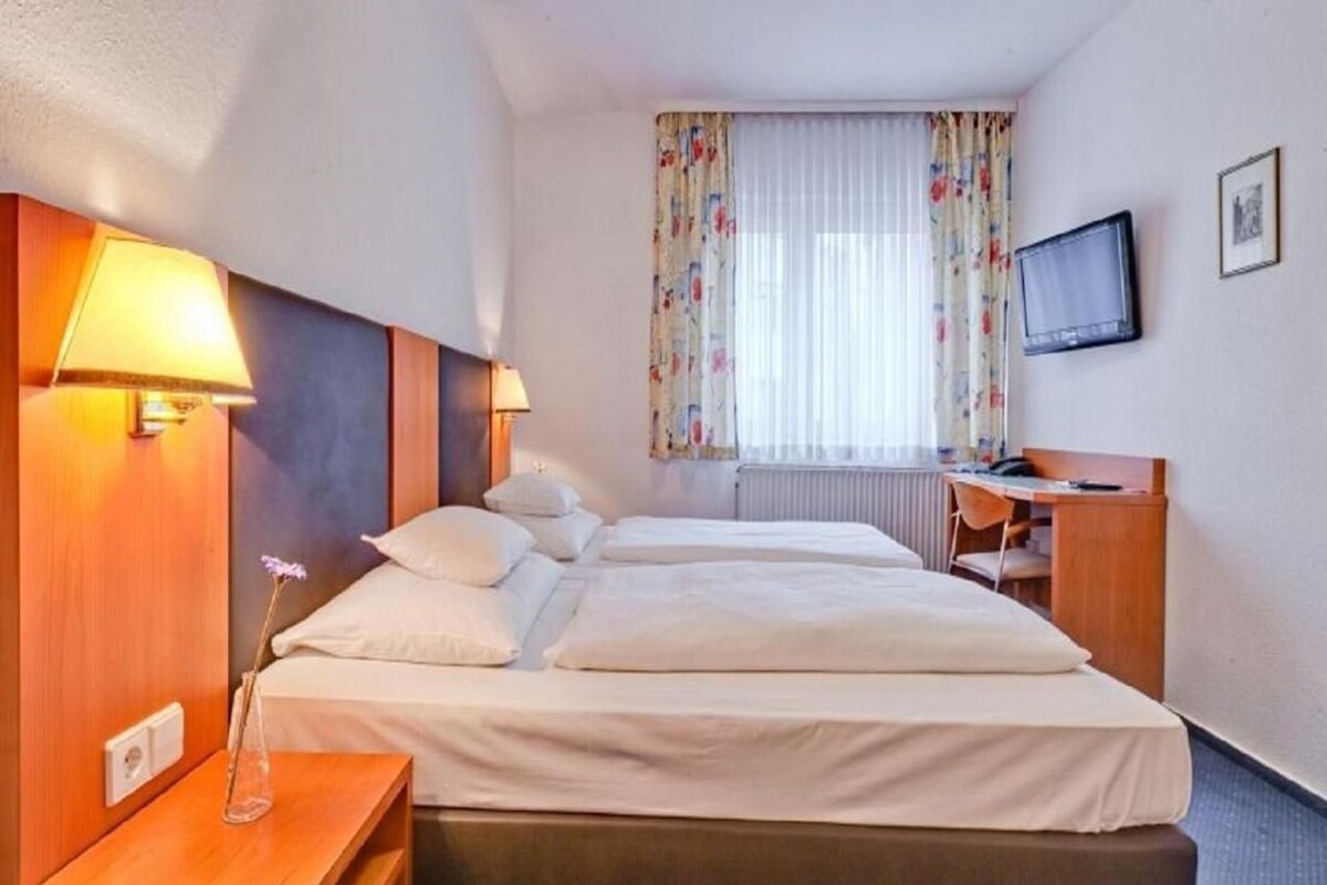 Habitación de hotel con cama doble, lámparas de noche, escritorio con silla, televisor y ventana con cortinas.