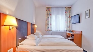 Hotellihuone, jossa on parivuode, yöpöytälamput, työpöytä tuolilla, televisio ja ikkuna, jossa on verhot.