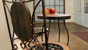 A chair with a metal frame and a wooden seat, a round table with a patterned top, and a window with a decorative grille in a room with wooden flooring and a curved wall. - Villa Arborea (Augsburg)