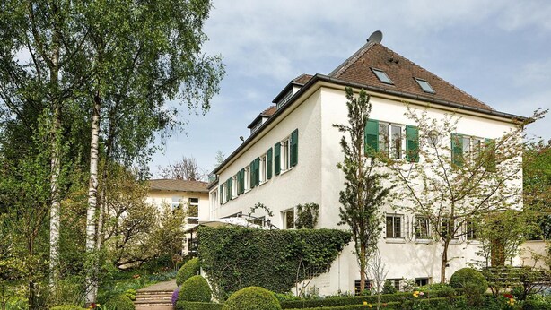 A two-story building with a white facade and green shutters, surrounded by well-maintained gardens and a cobblestone pathway. - Villa Arborea (Augsburg)