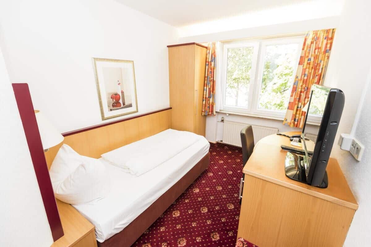 Quarto de hotel pequeno com uma cama de solteiro, uma escrivaninha com computador, janela com cortinas e um quadro na parede.