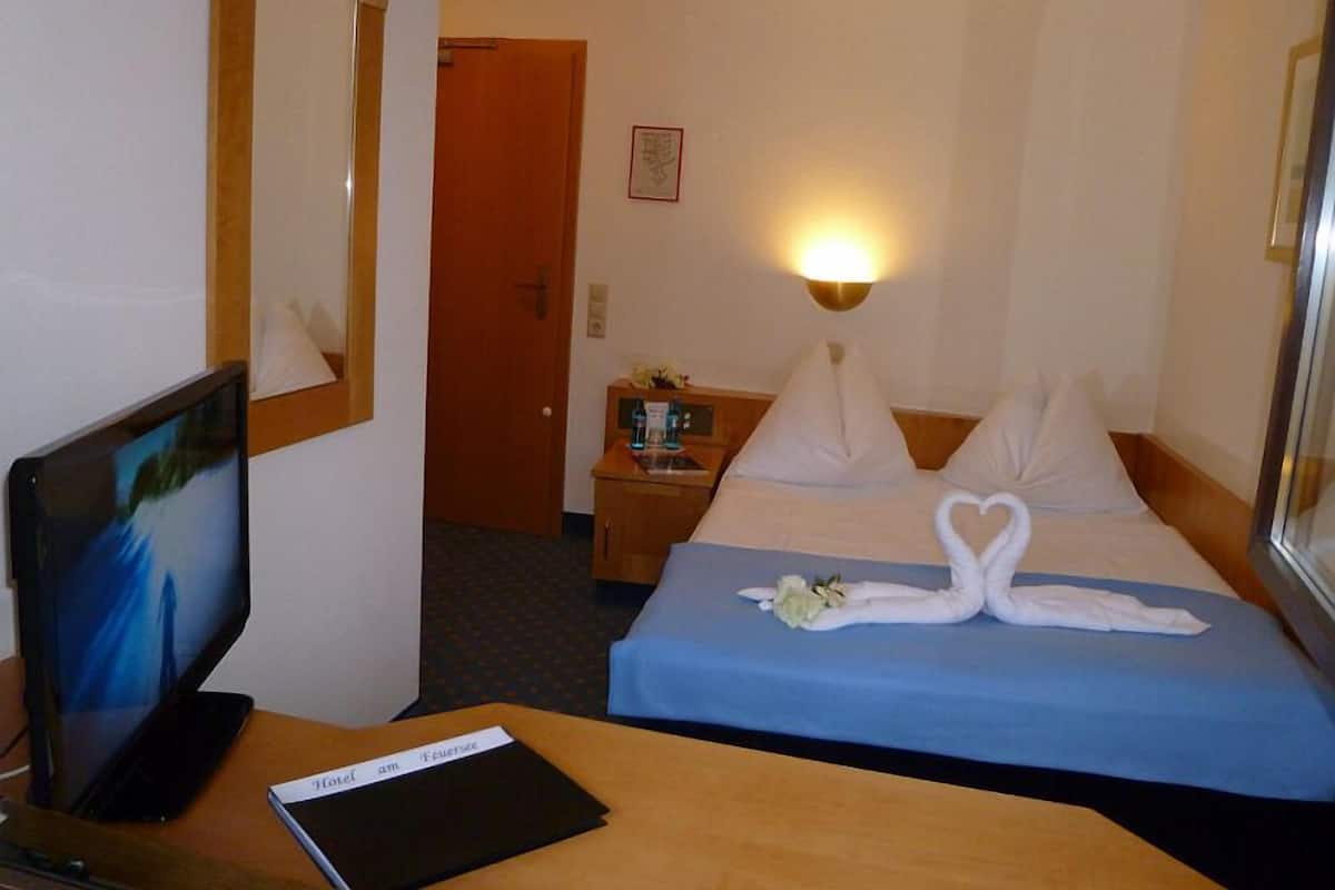 Quarto de hotel com uma cama, uma escrivaninha com computador, mesa de cabeceira com abajur e uma porta.
