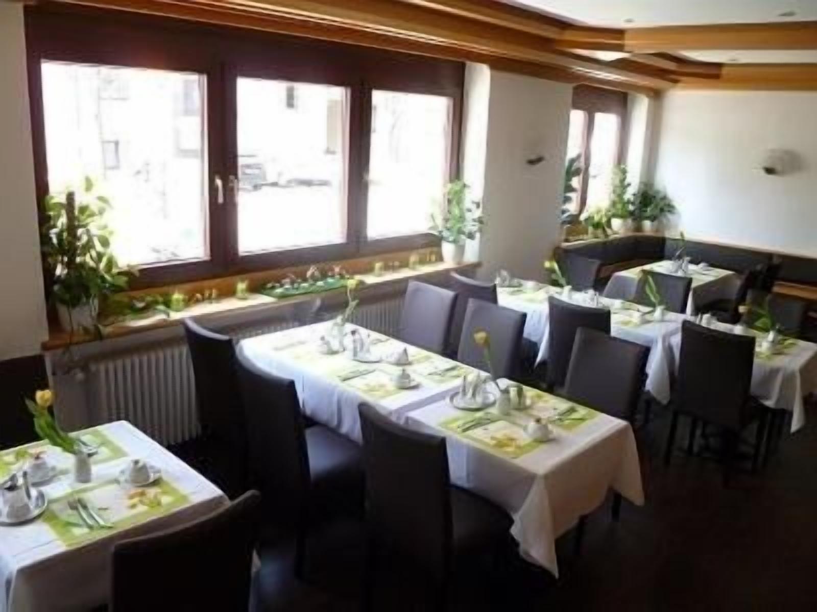 Un restaurant avec des tables dressées pour le service, de grandes fenêtres et des plantes d’intérieur.