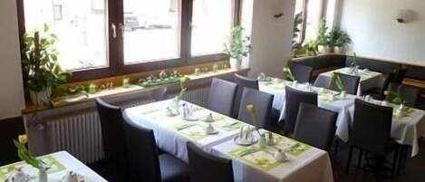 Un restaurant avec des tables dressées pour le service, de grandes fenêtres et des plantes d’intérieur.
