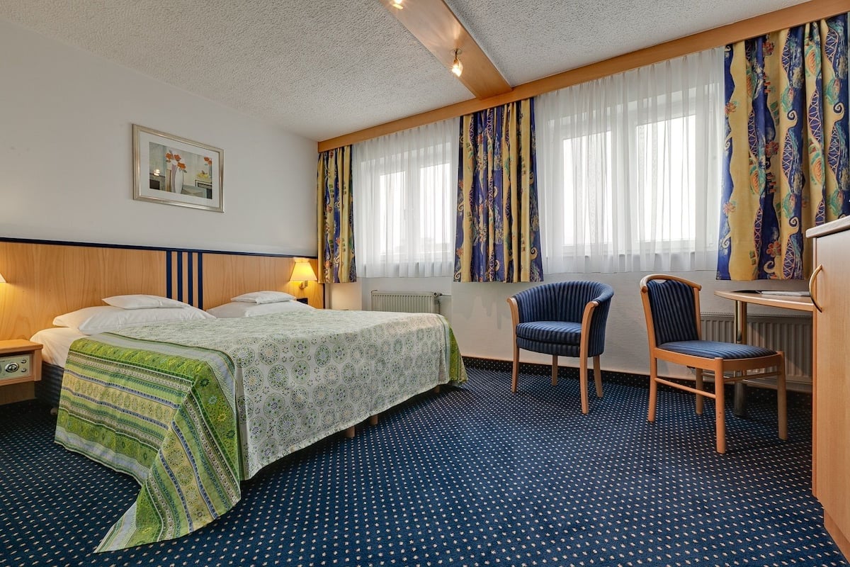 Quarto de hotel com uma cama, duas cadeiras, uma escrivaninha e uma janela com cortinas.