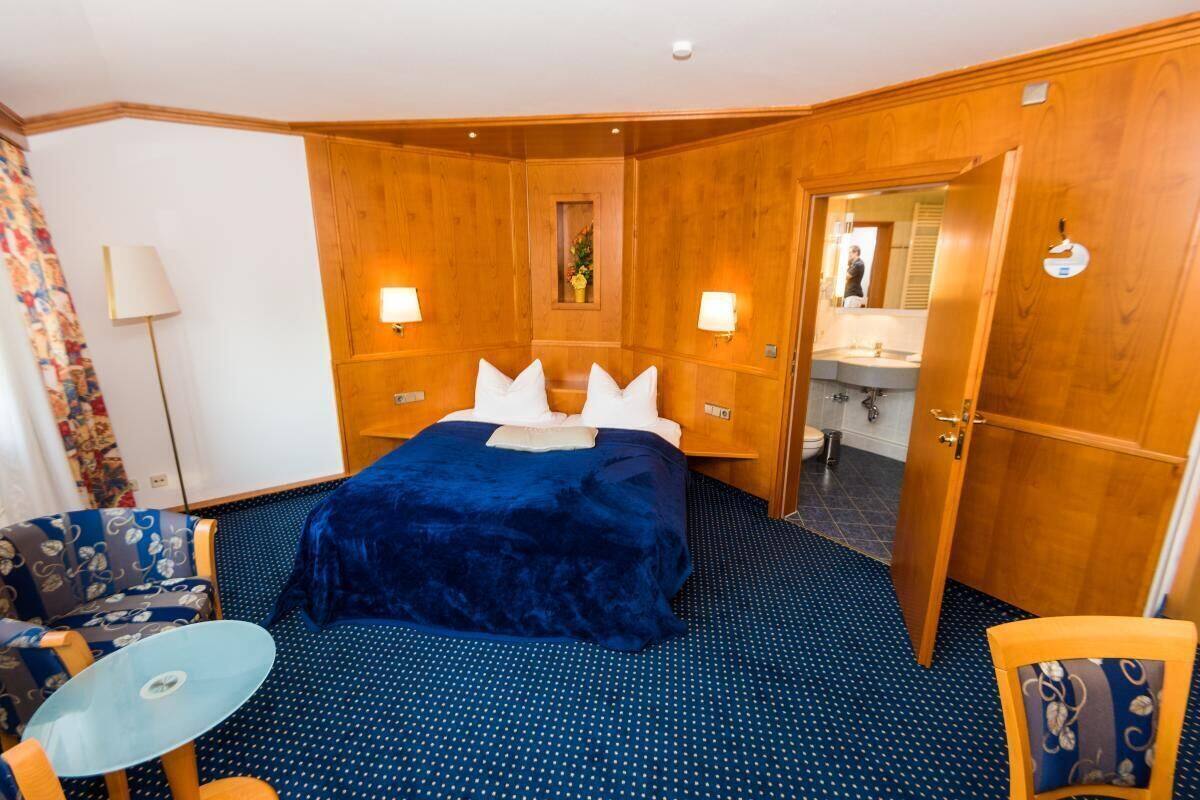 Quarto de hotel com uma cama grande, duas luminárias de cabeceira, uma mesinha, uma cadeira e um banheiro visível por uma porta aberta.