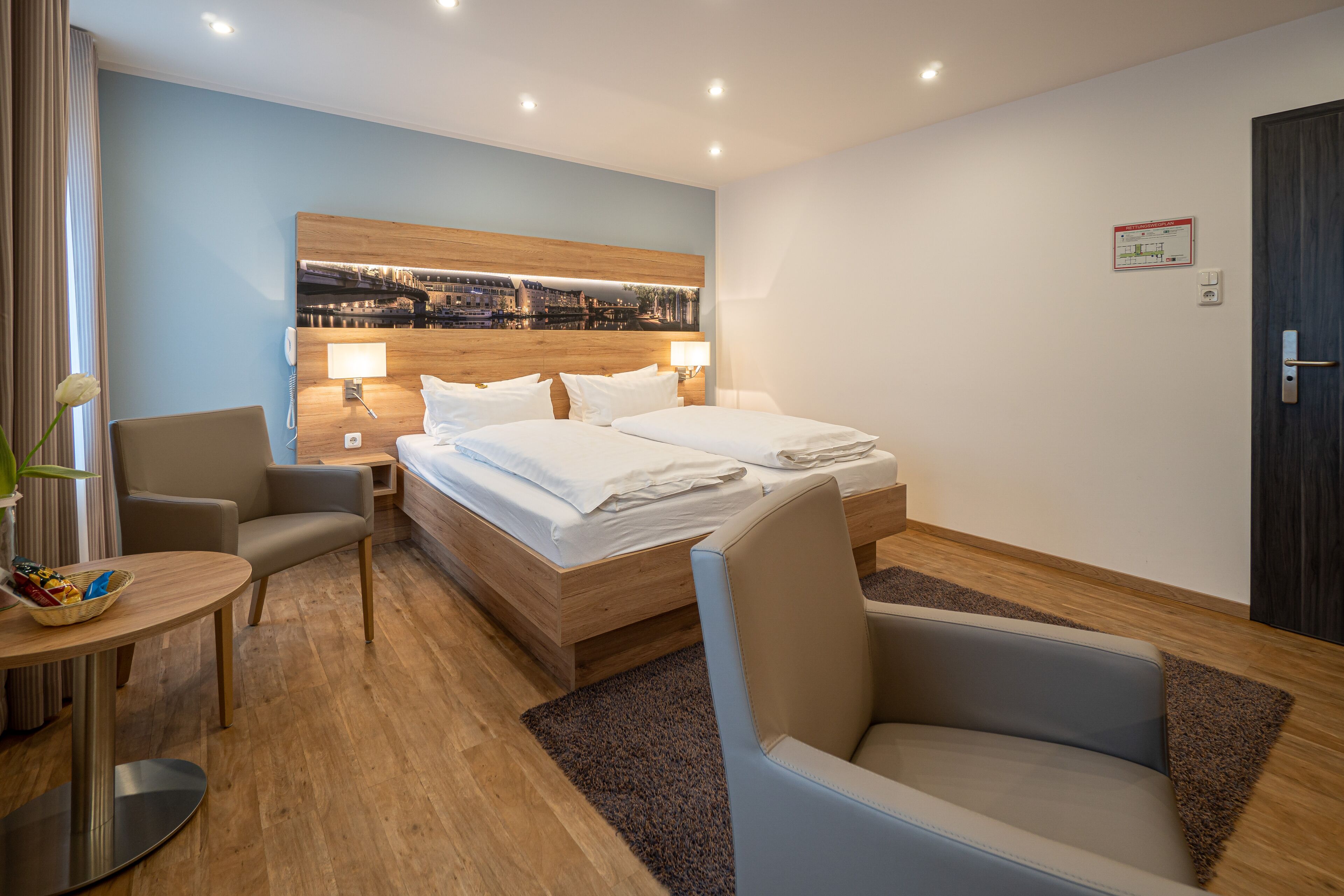 A hotel room with a bed, two bedside tables, a chair, a small table, and a cityscape artwork on the wall.
