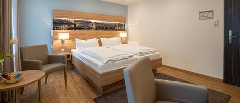 A hotel room with a bed, two bedside tables, a chair, a small table, and a cityscape artwork on the wall.