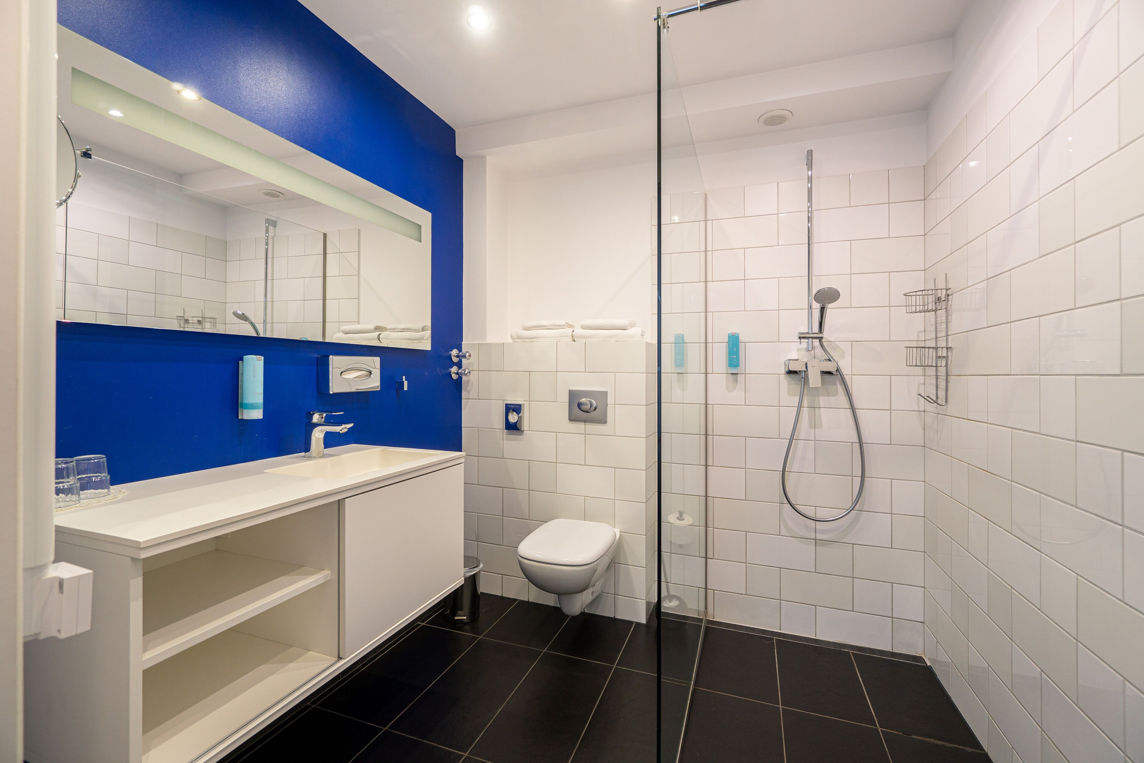 A modern bathroom with a blue accent wall, a white toilet, a glass-enclosed shower, and a white sink with a mirror above it.