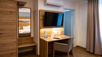 A compact hotel room with a wooden desk, a flat-screen TV, a mirror, and a bed with white linens.