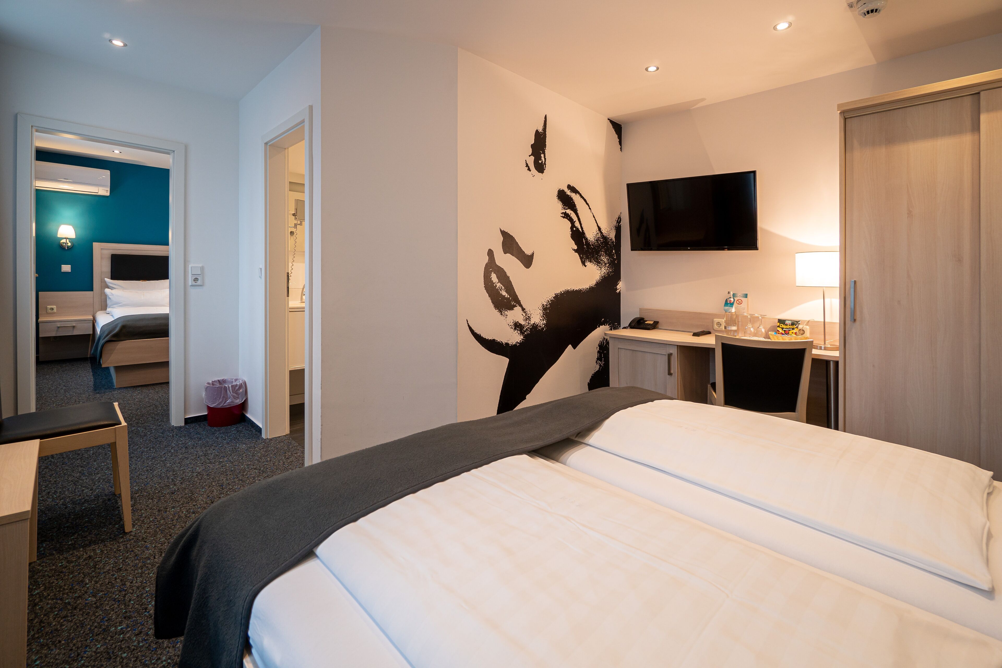 Ein Hotelzimmer mit einem Bett, einem Schreibtisch, einem Stuhl, einem Fernseher und einem durchgehenden Türspalte sichtbaren Badezimmer.