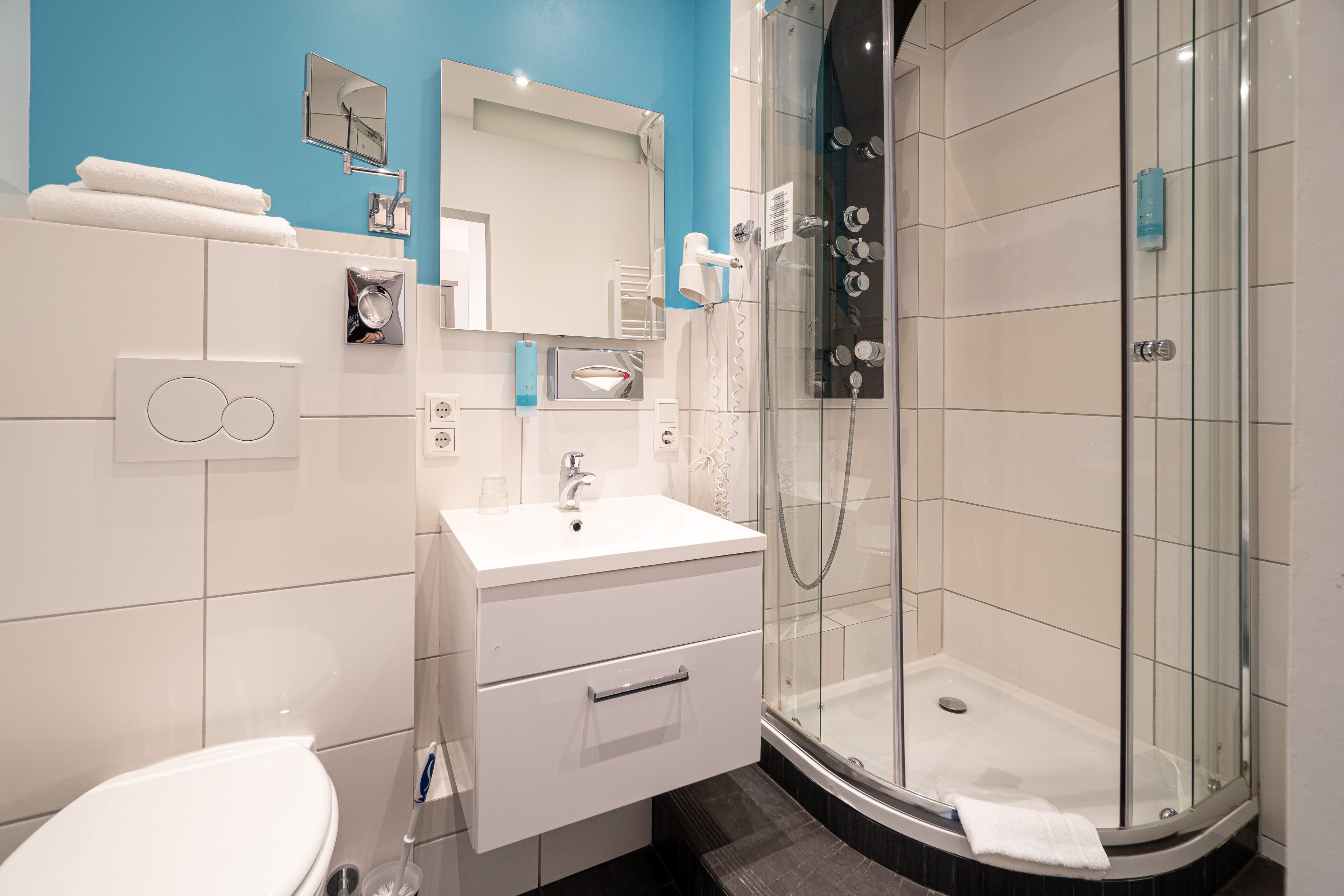 Un baño moderno con un lavamanos blanco, espejo e inodoro. Incluye una ducha con mampara de vidrio y un perchero para toallas.