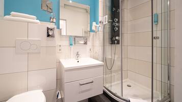 Un baño moderno con un lavamanos blanco, espejo e inodoro. Incluye una ducha con mampara de vidrio y un perchero para toallas.