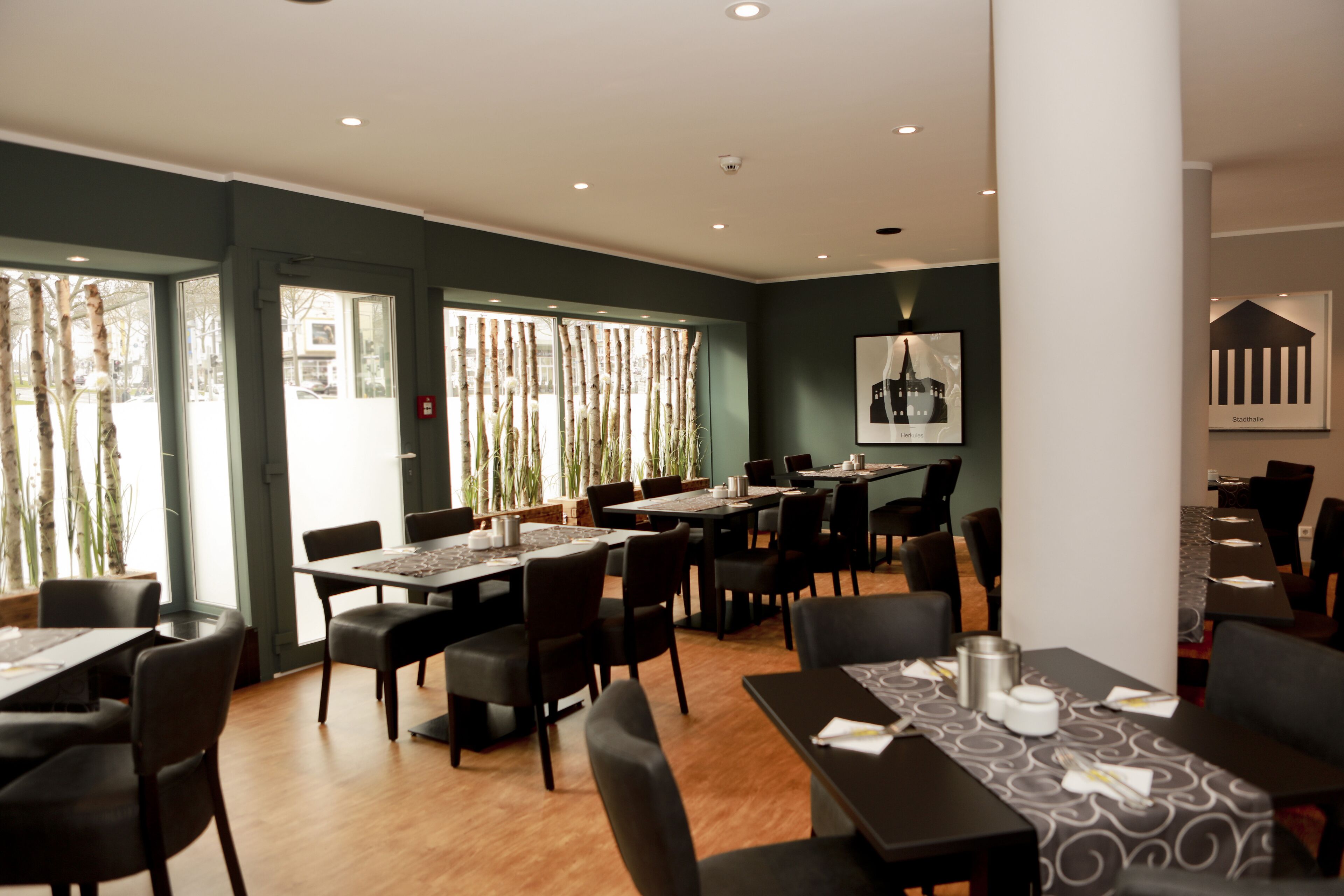 A modern dining area with multiple tables and chairs, large windows with natural light, and a framed picture on the wall.