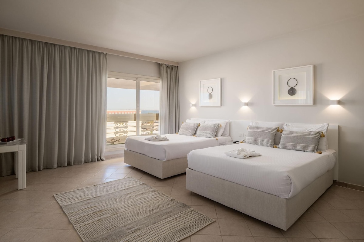 Habitació d'hotel amb dos llits, una finestra gran amb cortines, una taula petita i vistes a la platja.