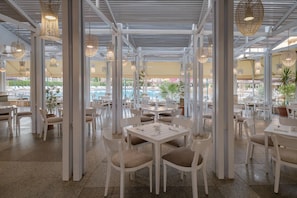 An indoor dining area with white tables and chairs, large windows, and hanging light fixtures. - Sindbad Club (Hurghada)