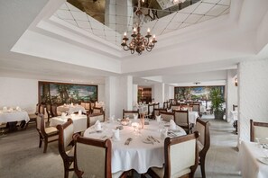 A spacious dining area with multiple tables set for dining, a chandelier, and a large painting on the wall. - Sindbad Club (Hurghada)