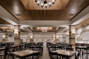 Un'ampia sala da pranzo con diversi tavoli e sedie, soffitto in legno con motivo geometrico e illuminazione a sospensione.