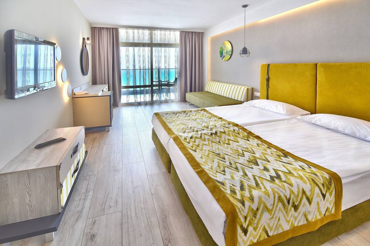 A hotel room with a bed, a desk, a chair, a large window with a view, and a balcony.