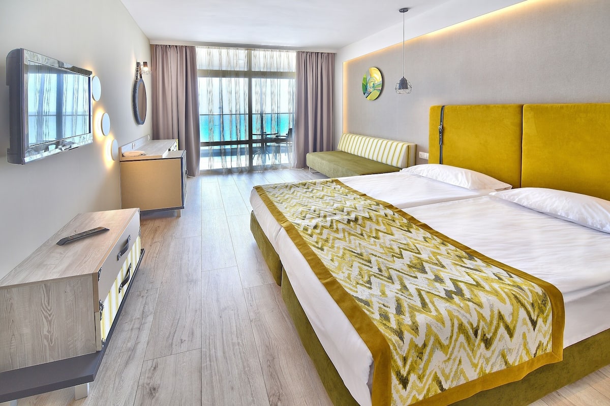 A hotel room with a bed, a desk, a chair, a large window with a view, and a balcony.
