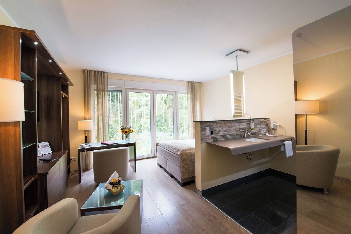 Habitación de hotel moderna con cama, escritorio con lámpara, silla, mesita con fruta, baño con lavabo y espejo, y ventanas amplias con vistas a árboles.