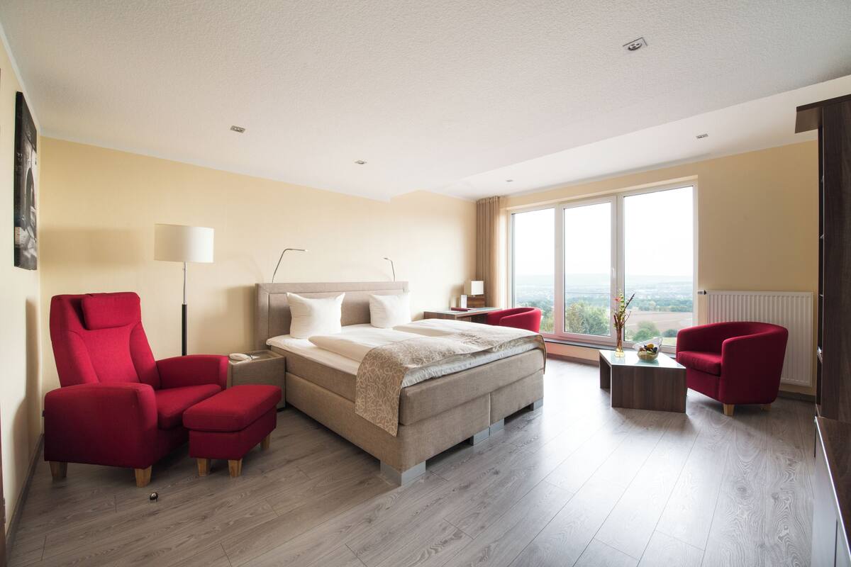 Una habitación de hotel moderna con una cama grande, dos sillones rojos, una mesita y vistas a la ciudad.